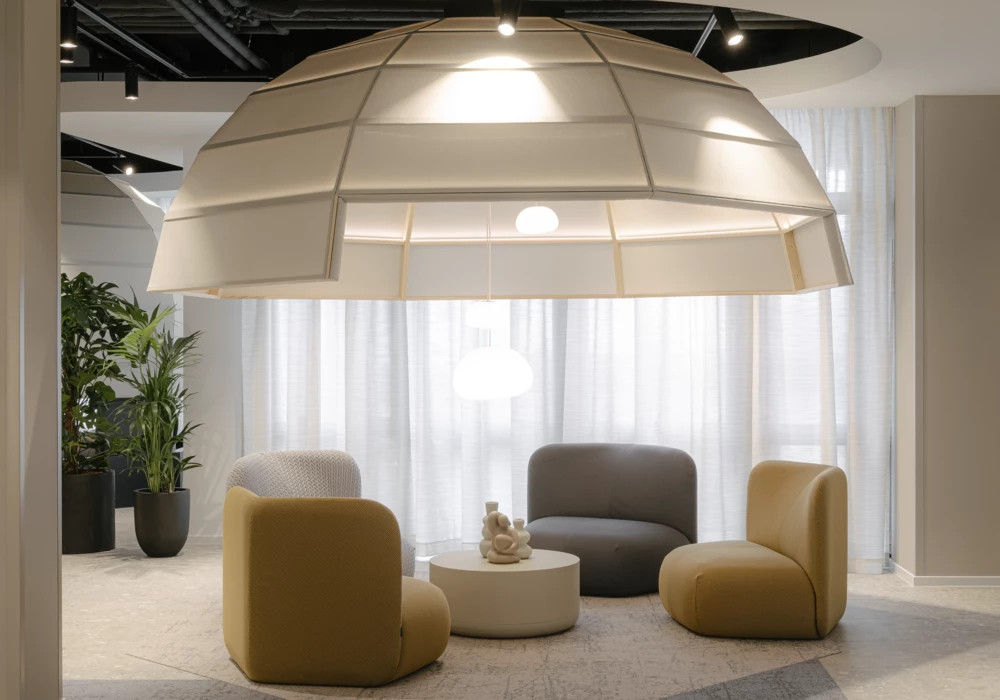 Modern lounge featuring a unique oversized light fixture resembling a floating dome. Beneath it, a seating arrangement includes upholstered chairs in beige, gray, and yellow, surrounding a round white coffee table. Sheer curtains and green plants add a welcoming and stylish ambiance.