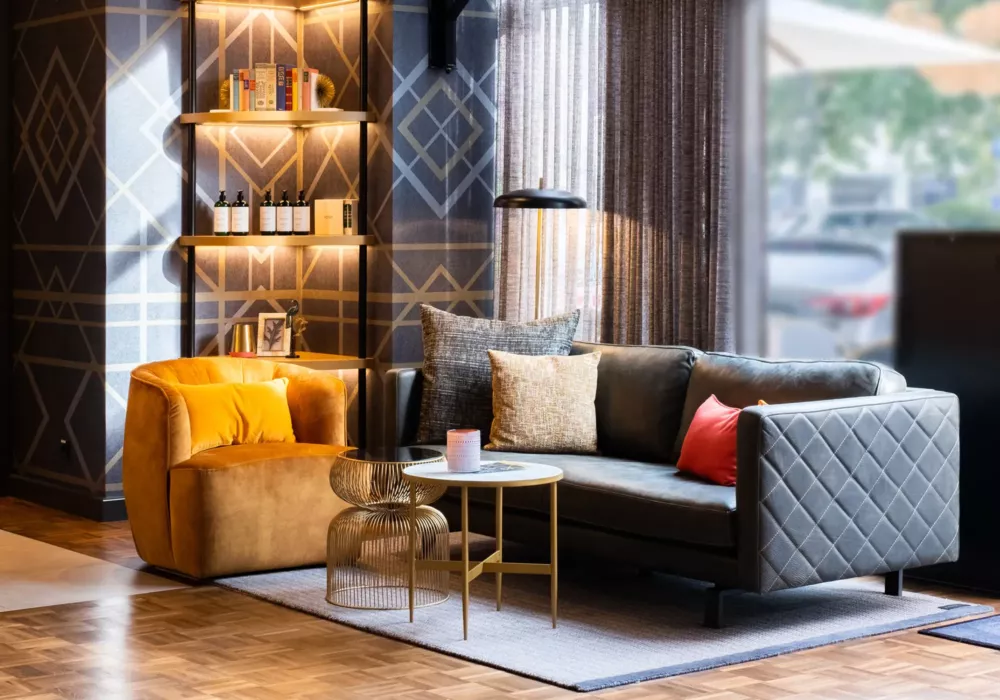 Stylish hotel lounge with a black leather sofa, mustard-yellow velvet armchair, gold side tables, and an illuminated shelf against a geometrically patterned wall.