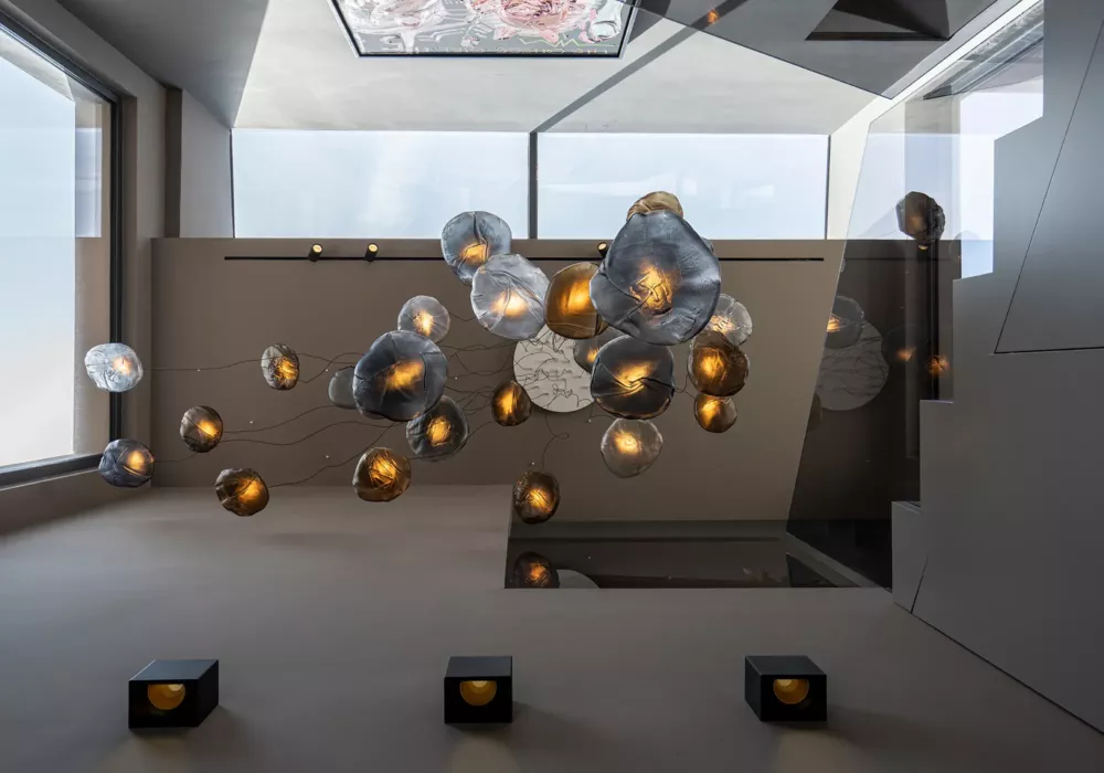 Modern designer pendant light with organically shaped, semi-transparent glass diffusers in gold and smoky gray tones. The sculptural lighting installation floats in a stylish, minimalist interior with large windows and mirrors.