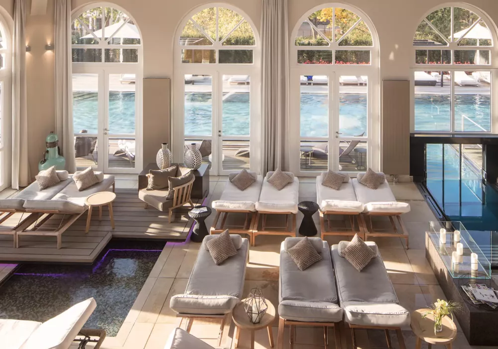 Elegant spa area at Schloss Hotel Fleesensee featuring stylish loungers, plush cushions, and a luxurious indoor pool. Large windows overlooking the outdoor pool create a bright and inviting ambiance.