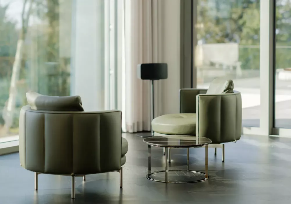Modern seating area with two green leather armchairs, a round glass table, and floor-to-ceiling windows with an outdoor view.