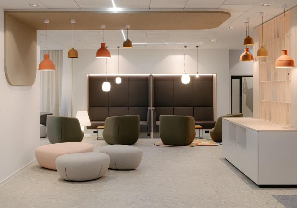 Modern lounge featuring a mix of upholstered seating poufs in beige and olive green, along with tall, dark-upholstered seating alcoves with integrated lighting. Multiple pendant lights in warm earth tones hang from the ceiling, creating a cozy atmosphere. A bright counter with wooden details completes the stylish design.