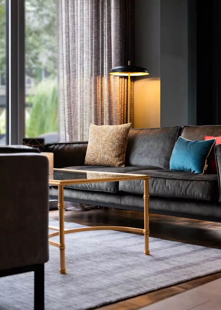 Elegant hotel lounge with a black leather sofa, decorative cushions, a gold coffee table, and ambient lighting in front of floor-to-ceiling windows.