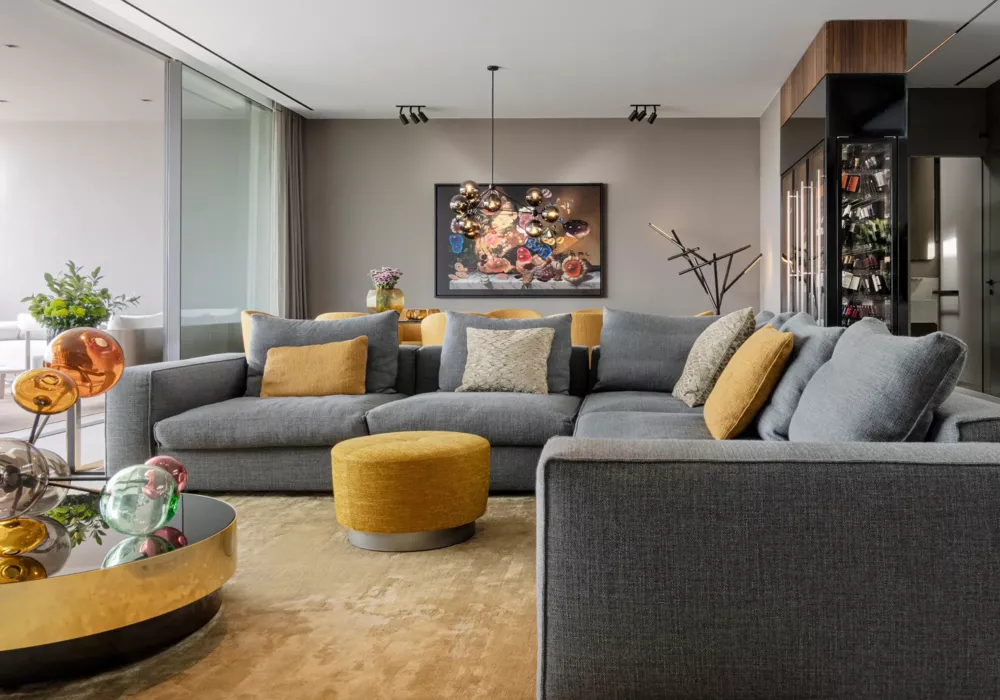 Modern living room with a large gray sectional sofa, golden yellow accents, and a sculptural designer lamp. A round coffee table with colorful glass spheres and a sleek wine cabinet complete the stylish interior.