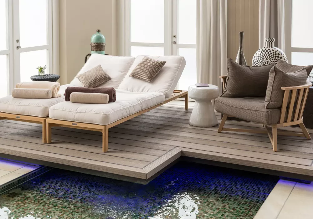 Two luxurious loungers with white cushions and pillows on a raised wooden platform above a water basin, next to a cozy armchair with a side table in a stylish spa area.