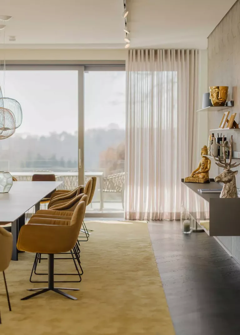 Elegant dining room with a long table, mustard-colored chairs, and large windows overlooking the terrace. Decorative shelves with golden sculptures adorn the wall.