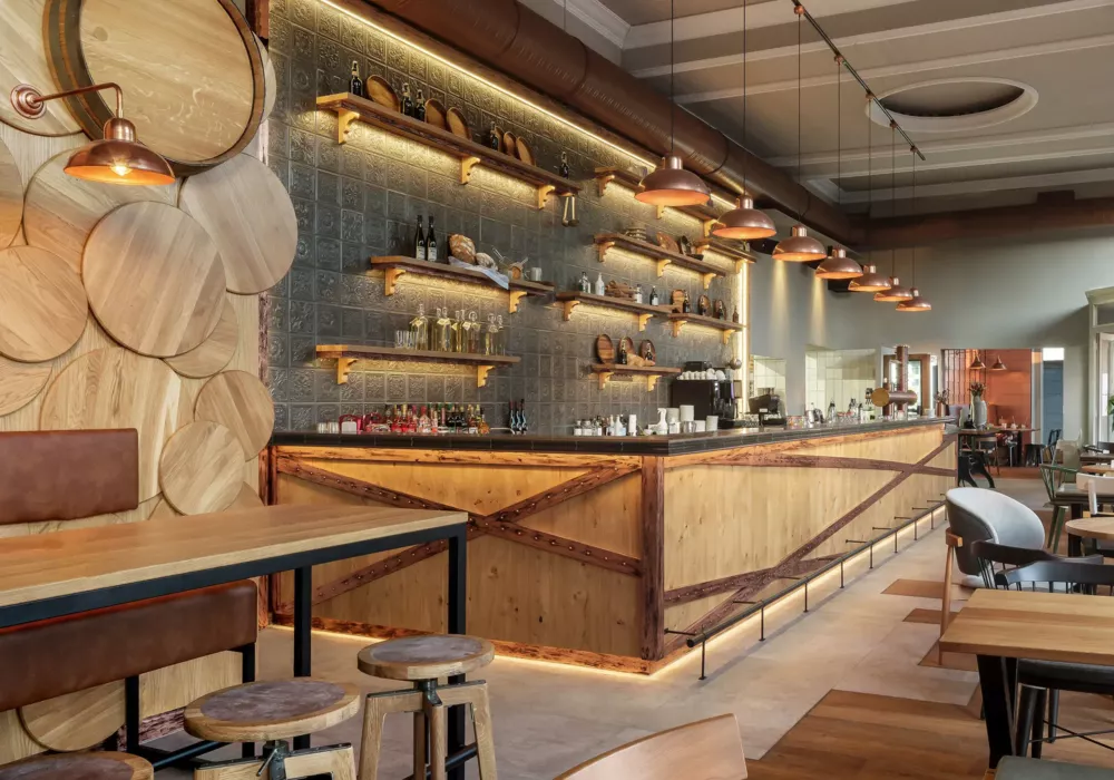 Modern tavern with a long wooden bar counter, warmly lit shelves with beverages, and rustic wooden furniture in an industrial design.