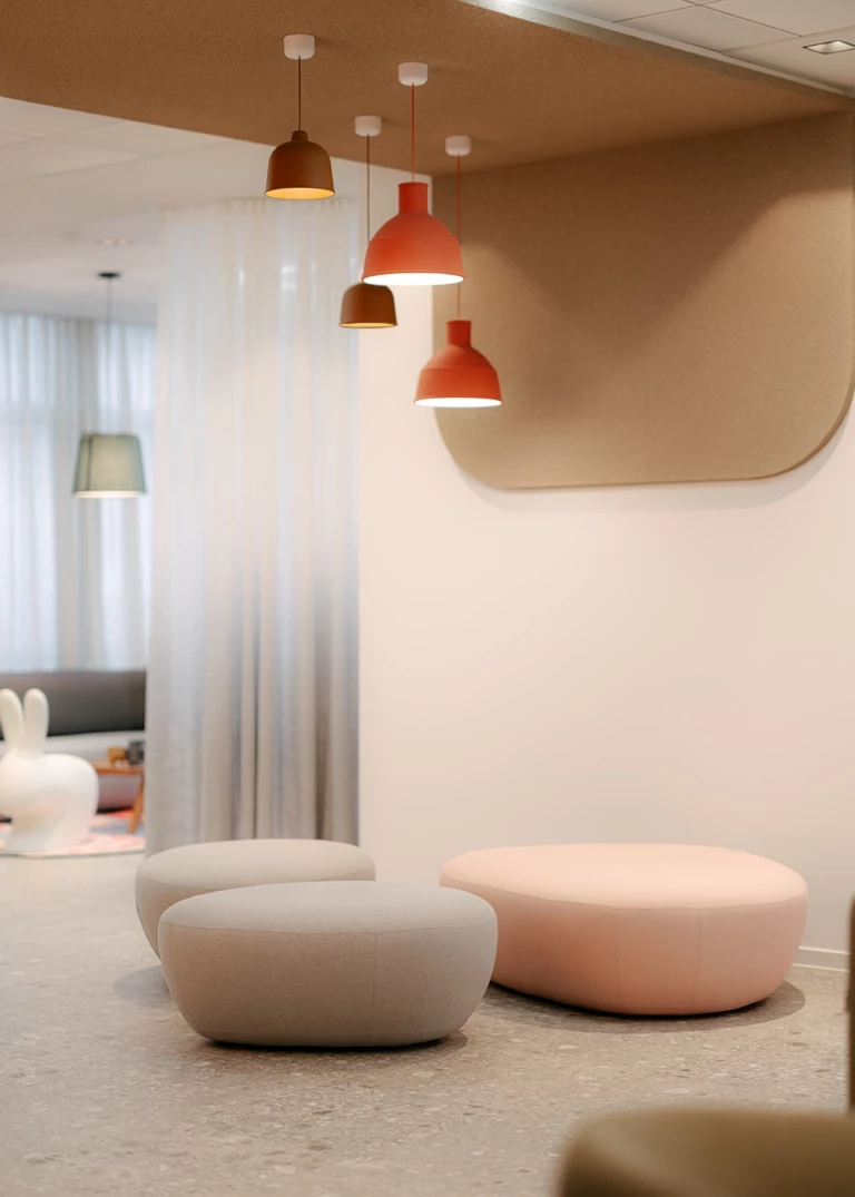 Modern lounge featuring round upholstered poufs in beige and pink on a light-colored floor. Above them, multiple pendant lights in warm earth tones hang from the ceiling. In the background, semi-transparent curtains create a soft lighting atmosphere, while a playful rabbit decor element adds a unique touch.