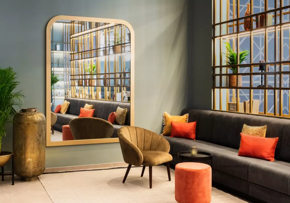 Stylish hotel lounge with a black upholstered bench, golden-brown armchair, colorful cushions, and decorative accents. A large mirror reflects the modern shelving unit with gold elements.