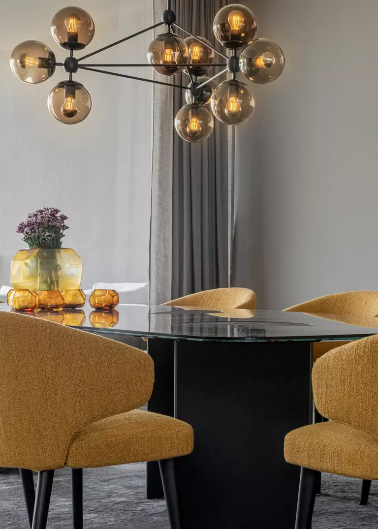 Elegant dining room with a glass dining table, golden yellow upholstered chairs, and a modern chandelier with smoked glass globes. Decorative yellow vases with flowers add stylish accents.