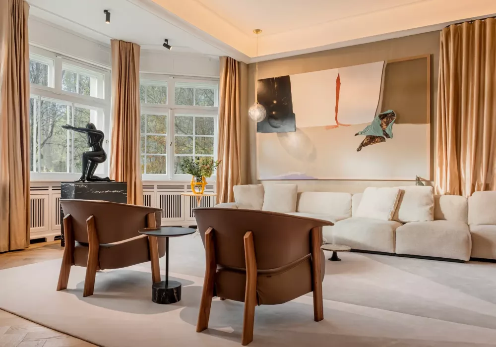 Stylish living room featuring a large white sofa, two brown leather armchairs, and an abstract artwork on the wall. A black sculpture on a marble pedestal and golden curtains add elegance to the space.