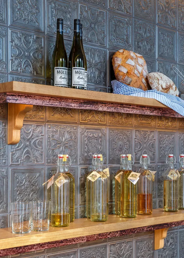 Wooden shelves with wine bottles, homemade beverages in glass bottles, and freshly baked bread against a decorative metal wall.