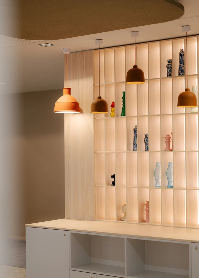 Modern light wood shelving unit with integrated lighting, displaying decorative vases and sculptures. Above, multiple pendant lights in warm earth tones provide soft illumination. In the foreground, a white sideboard with open compartments completes the setting.