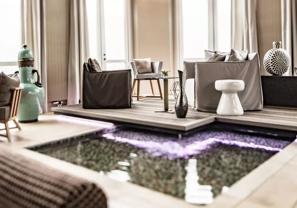 Elegant spa area with elevated seating on a wooden platform above an illuminated water basin, stylish armchairs, decorative vases, and modern furniture.