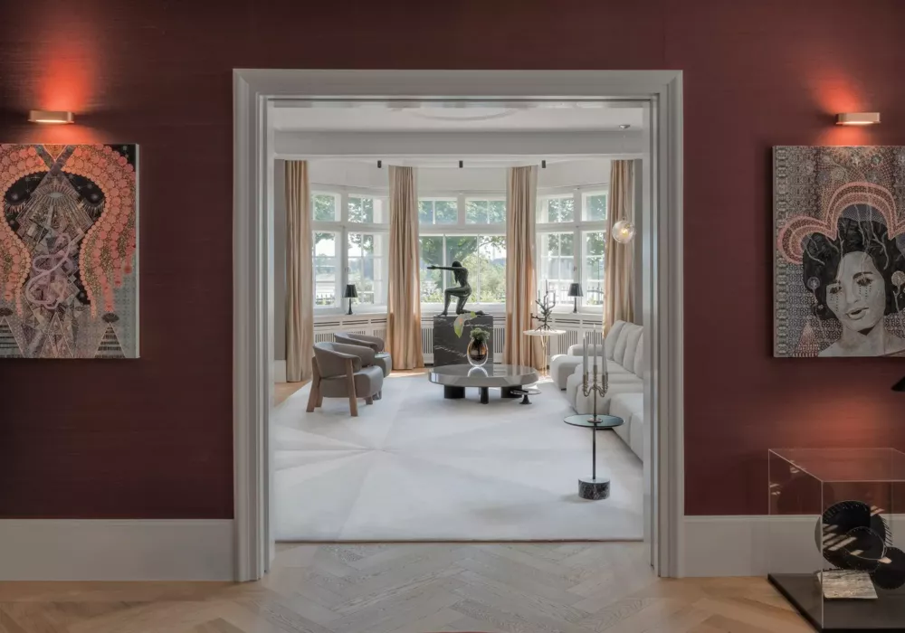 View through a wide doorway into an elegant living room with a white sofa, round coffee tables, and golden curtains. The deep red walls of the adjacent room feature two artistic portraits, illuminated by stylish wall lights.