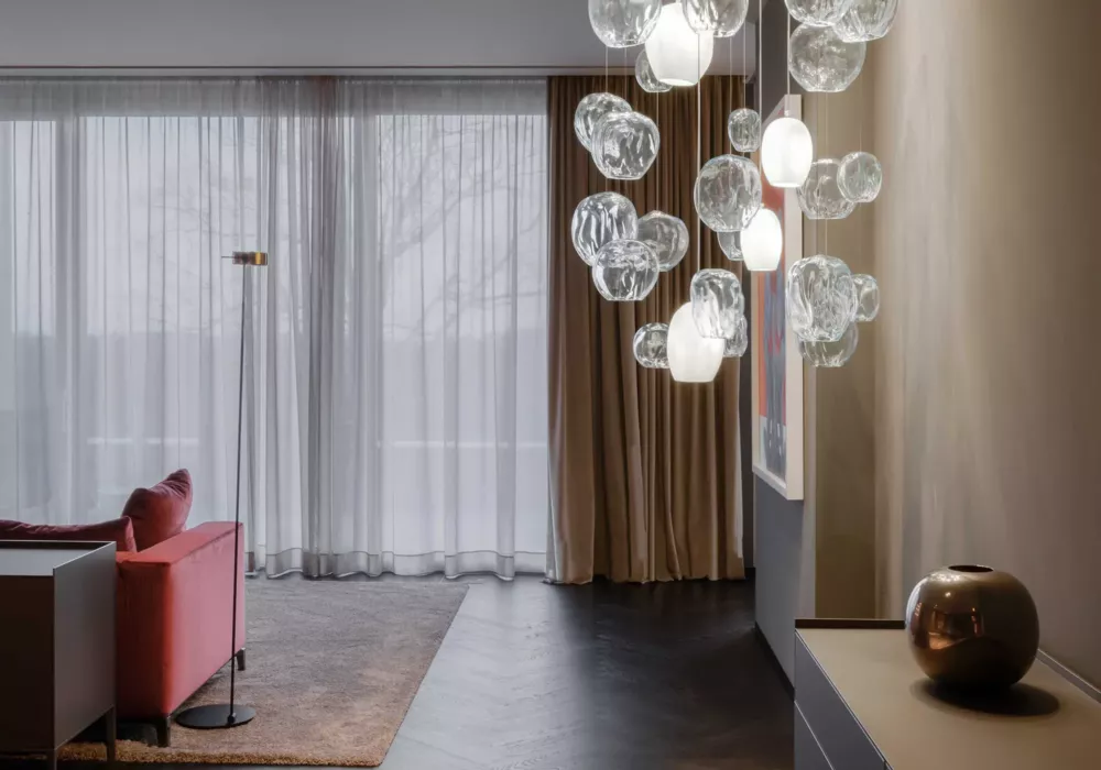 Modern living room with a red sofa, a sleek floor lamp, and a floor-to-ceiling window with sheer curtains. A striking glass pendant light hangs from the ceiling, while a bronze-colored vase sits on a console table.