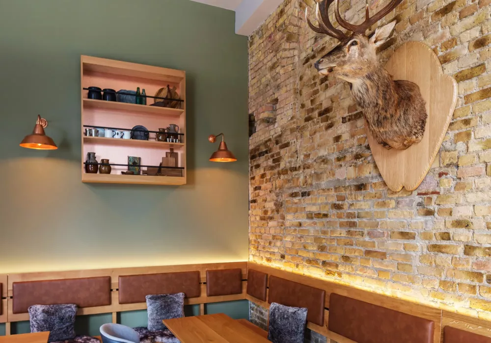 Rustic restaurant interior with an exposed brick wall, a mounted deer head, wooden shelves with ceramics, and copper wall lamps.