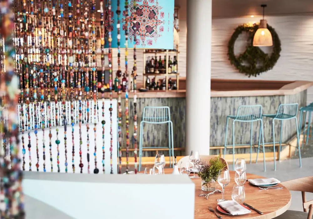 Stylish restaurant at 7Pines Resort Ibiza featuring a wooden dining table, colorful bead curtain, and a modern bar in the background