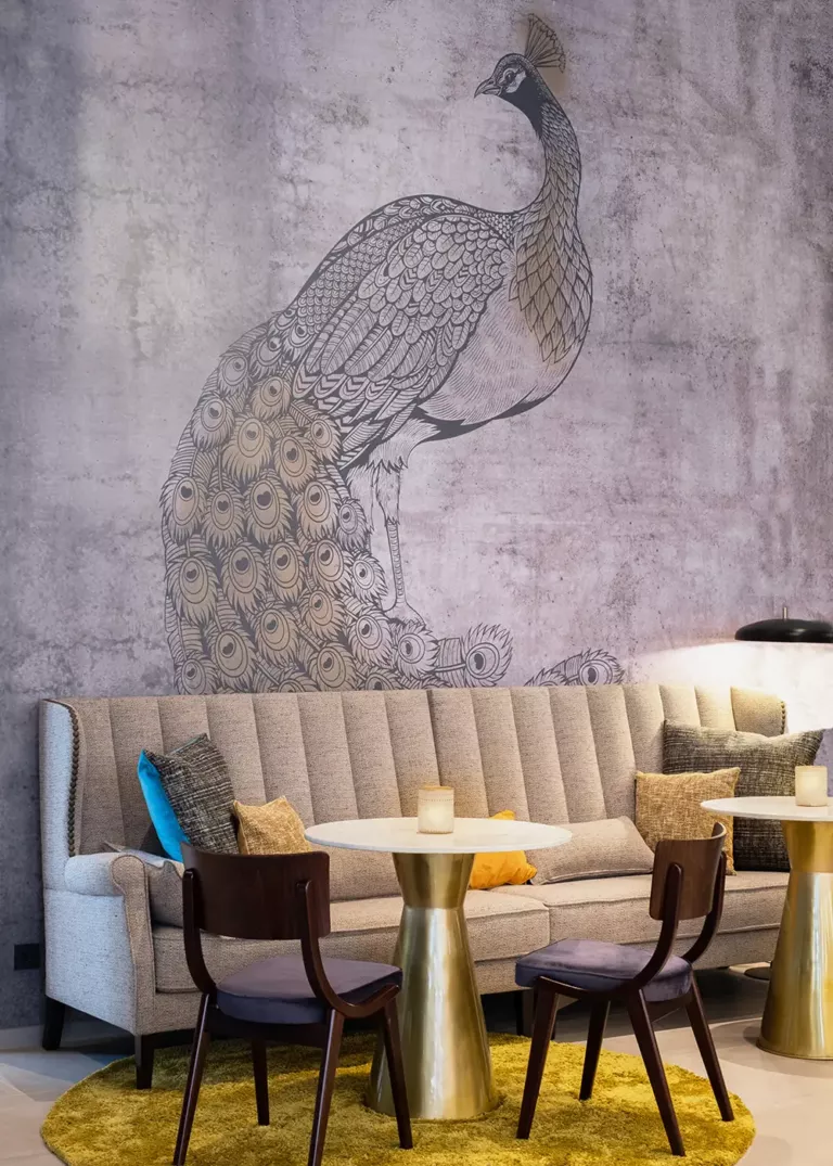 Stylish hotel lounge with a tall upholstered bench, round marble tables with gold bases, and dark wooden chairs. The wall design features an artistic peacock illustration on a textured surface.