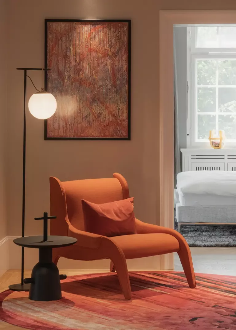 Cozy reading corner with an orange armchair, matching pillow, and a modern side table next to a floor lamp with a spherical light. An abstract artwork hangs on the wall, while a bedroom with a white bed is visible in the background.