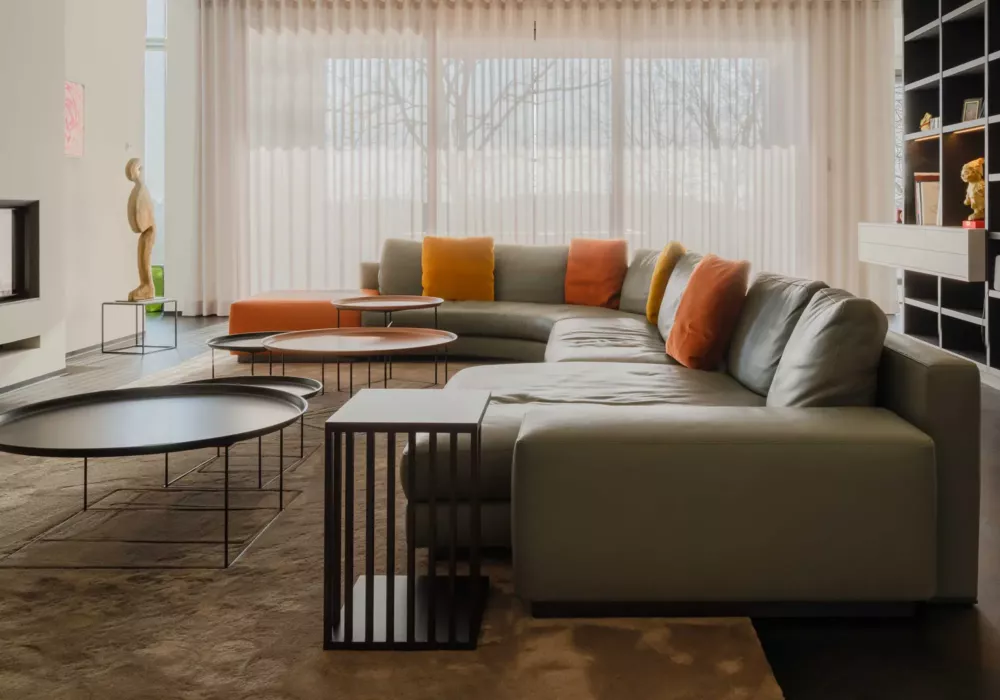Modern living room with a large gray sectional sofa, colorful cushions, multiple round coffee tables, and floor-to-ceiling windows with curtains.