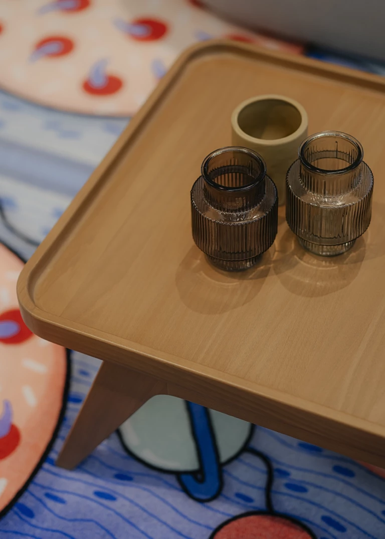 Close-up of a wooden side table with two ribbed glass vases in brown tones and a small ceramic container. In the background, a colorful rug with a playful pattern in blue, red, and beige.