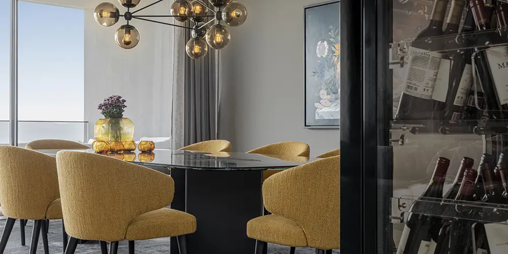 Modern dining room with a black glass table, mustard-yellow upholstered chairs, stylish chandelier, and a wine cabinet in the foreground.