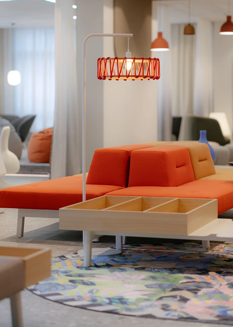 Modern lounge furniture with orange cushions and an integrated wooden table with compartments. A striking red pendant lamp complements the design. In the background, additional seating and a playful room layout are visible.