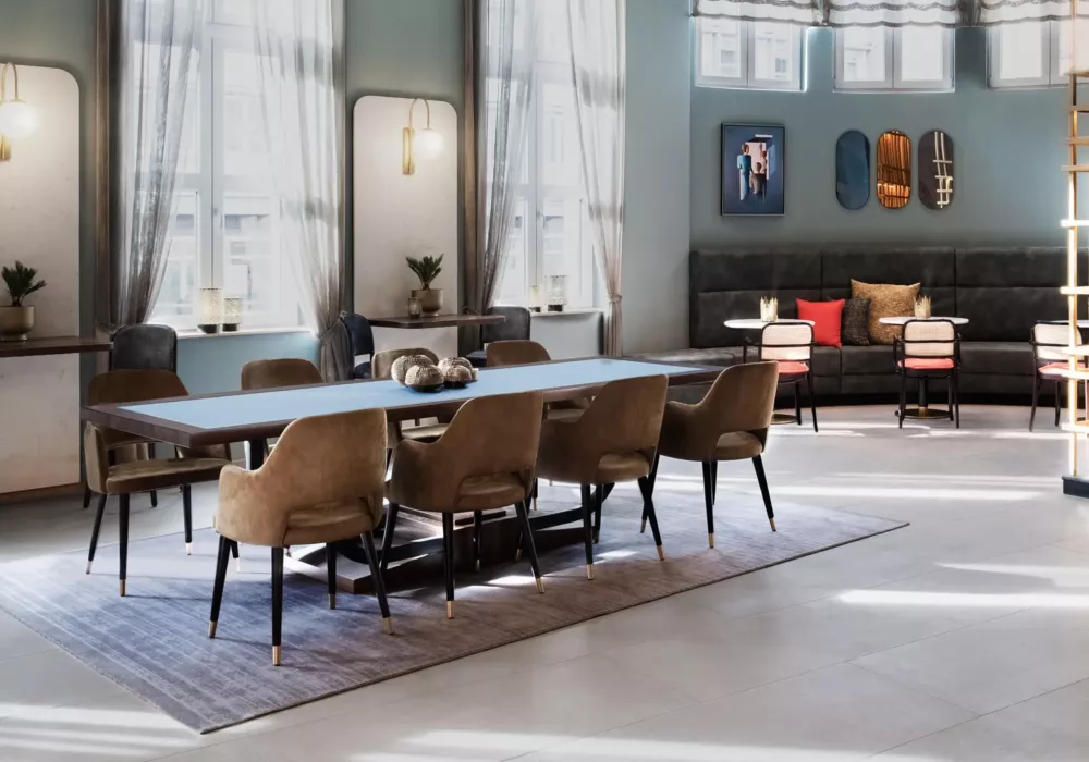 Modern hotel lounge featuring a long dining table surrounded by elegant beige velvet chairs with black legs and gold accents. A cozy seating area with a dark upholstered bench and decorative pillows adds to the ambiance. Large windows with sheer curtains create a bright and inviting atmosphere.