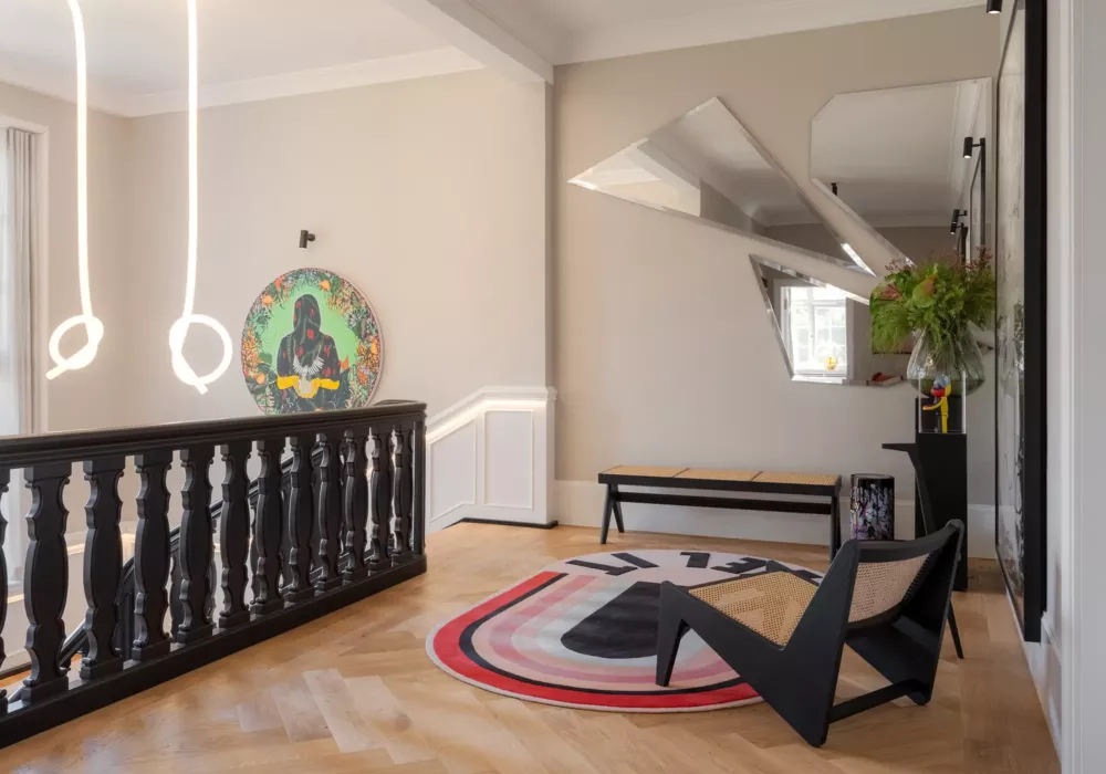 Modern hallway with a black wooden railing, designer chair, and a rug featuring the words Feel It. A round artwork with a colorful motif hangs on the wall, while large mirrors add an elegant touch. A stylish bench and a vase with flowers complete the interior.