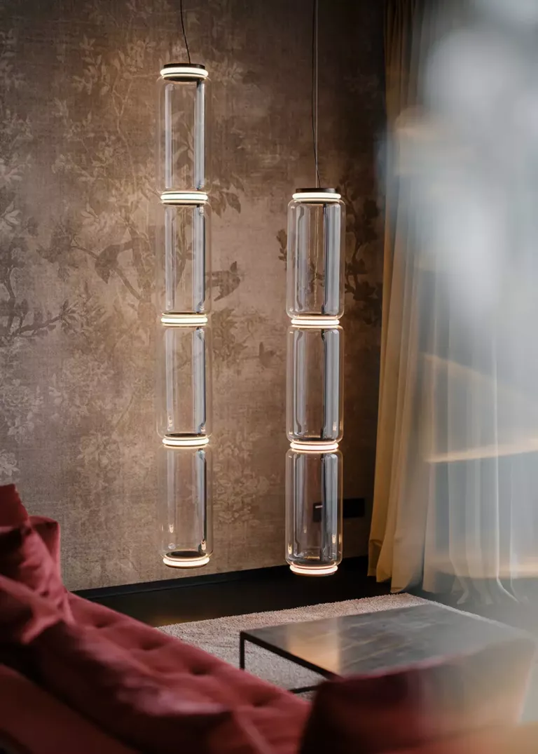 Elegant glass pendant lights with a cylindrical design, hanging in front of patterned wallpaper, next to a red sofa and a metal coffee table.