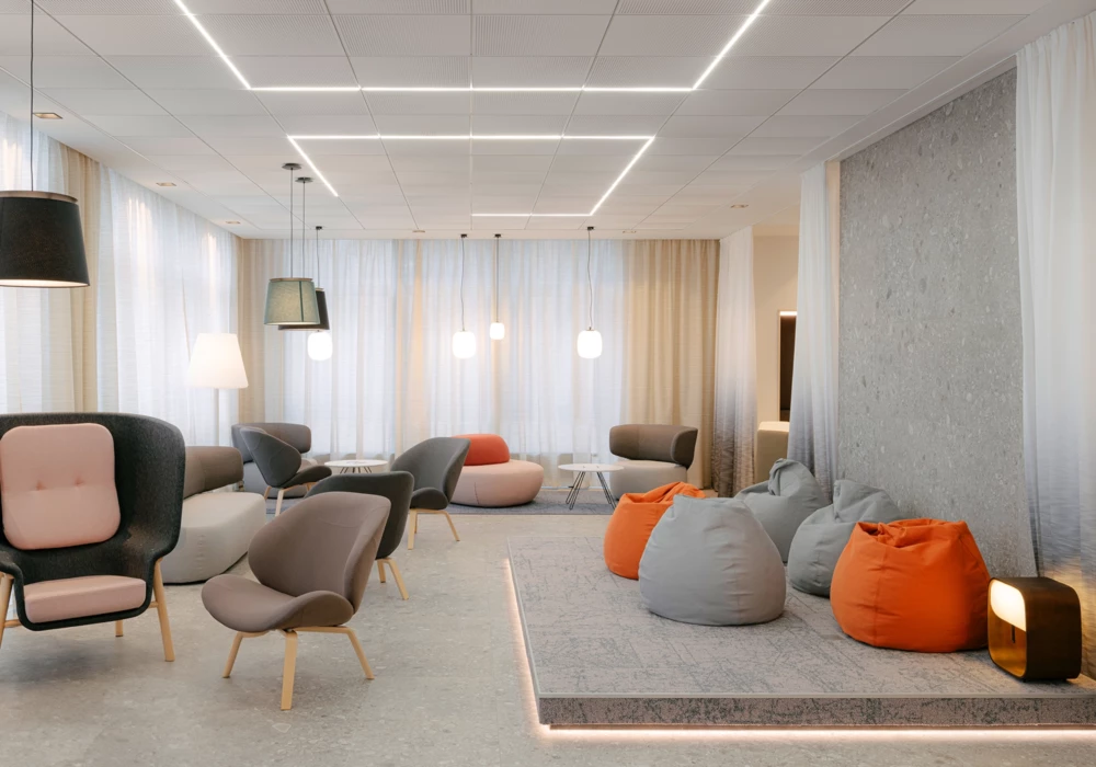 Modern lounge area with a mix of upholstered armchairs, bean bags in gray and orange, and minimalist lighting. Large windows with sheer curtains allow plenty of natural light into the space.