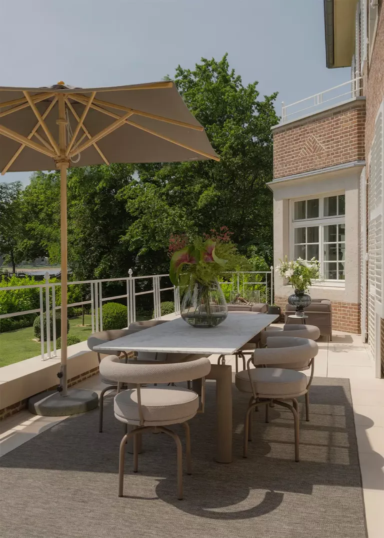 Stylishly designed terrace featuring a large stone dining table surrounded by modern upholstered chairs. A sun umbrella provides shade, while a vase with fresh flowers decorates the table. The terrace overlooks a well-manicured garden with lush trees and shrubs.