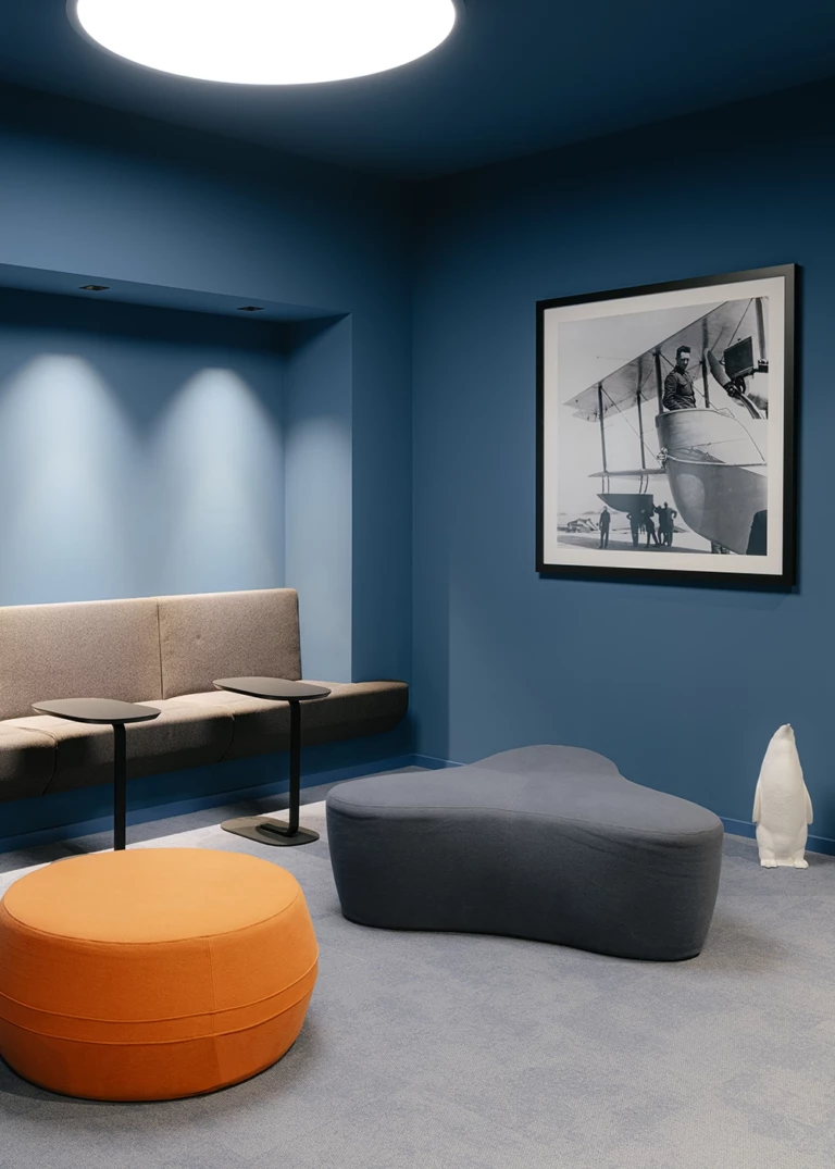 Modern lounge area with deep blue walls, a padded bench, and stylish stools in orange and gray. A black-and-white photo print of a historic airplane complements the design.