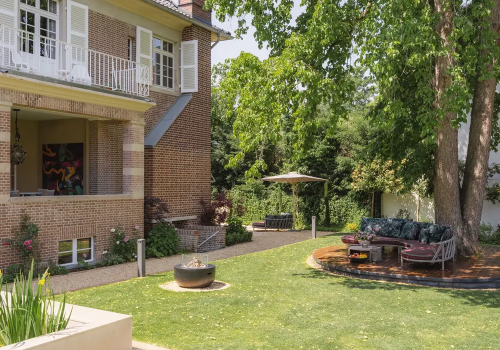 Elegant garden area featuring a stylish brick villa, surrounded by lush trees and a well-maintained lawn. A round lounge seating area with plush cushions sits on a wooden platform beneath a large tree. In the background, another seating arrangement with a sun umbrella provides a shaded retreat.