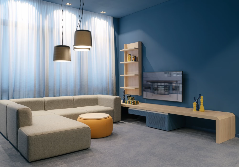 Stylish lounge with deep blue walls, a gray corner sofa, and a yellow ottoman as an accent. Wooden shelves and a modern TV wall complement the minimalist design.