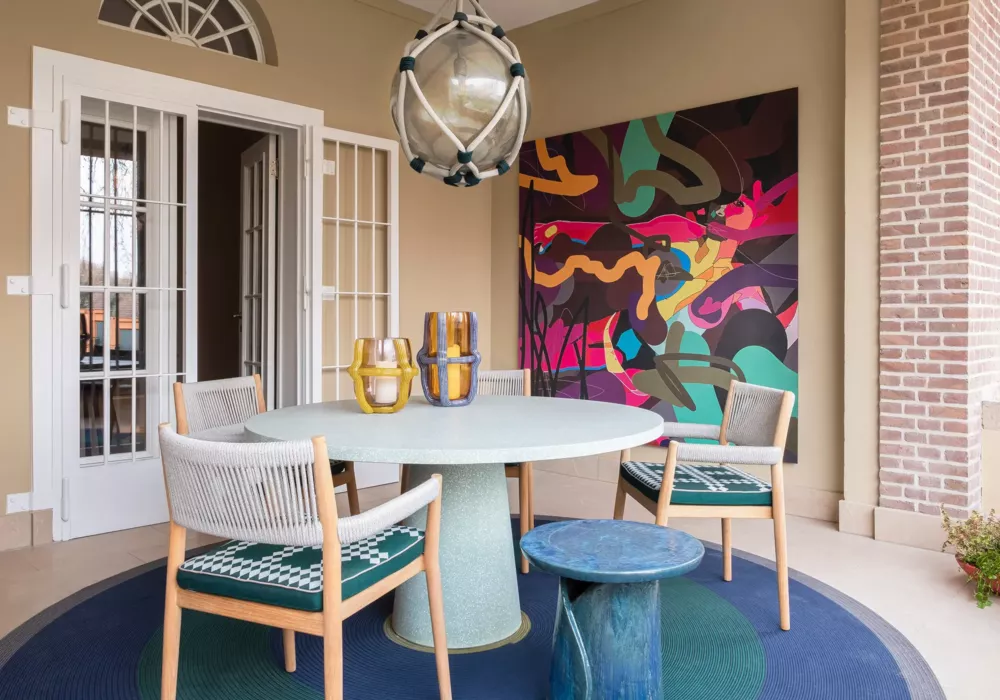 Stylish covered terrace featuring a round dining table and four modern chairs with woven seats. A colorful abstract painting adorns the wall, while decorative glass vases add accents to the table. A striking pendant light enhances the elegant atmosphere.