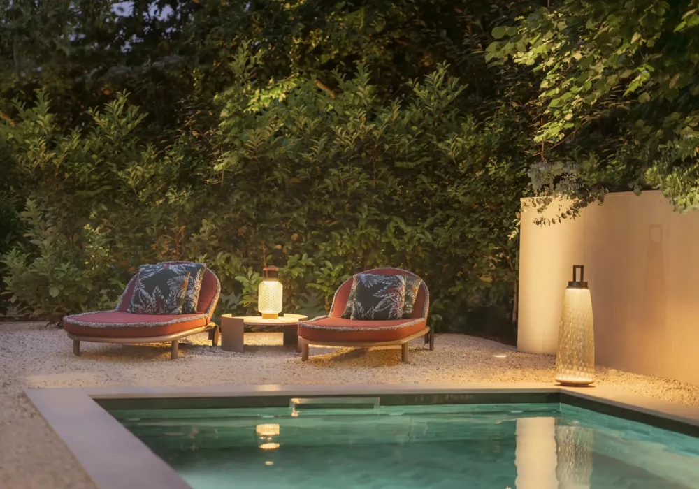 Stylish pool lounge at dusk with two round lounge chairs featuring red cushions and tropical-patterned pillows. A lantern and a side table sit between the chairs. The pool water is visible in the foreground.