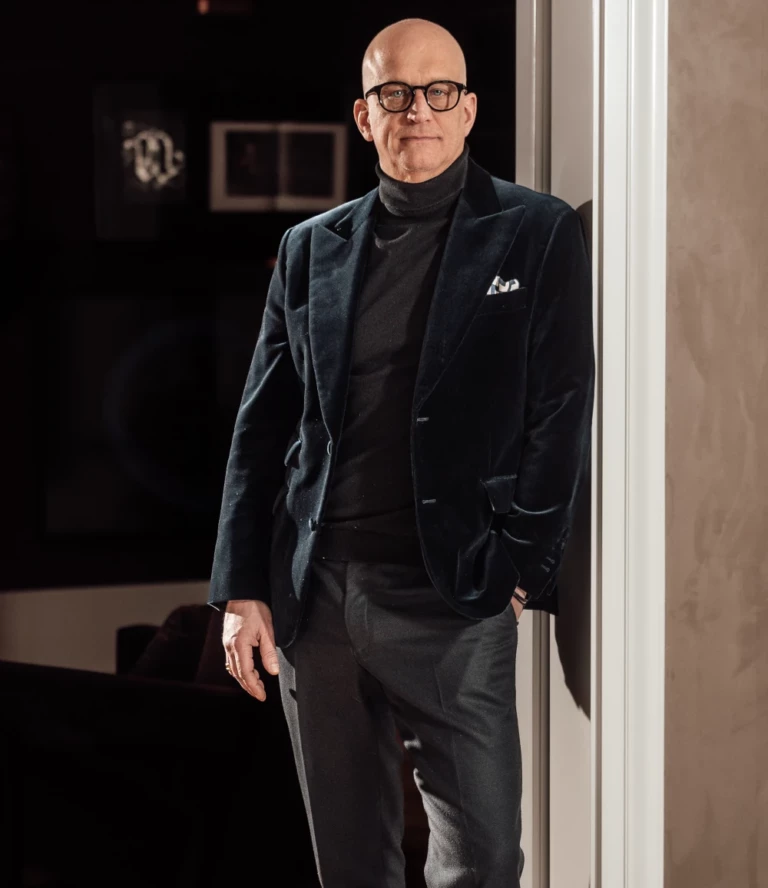 „Olaf Kitzig in eleganter dunkler Kleidung, stehend in einem stilvoll beleuchteten Innenraum.
