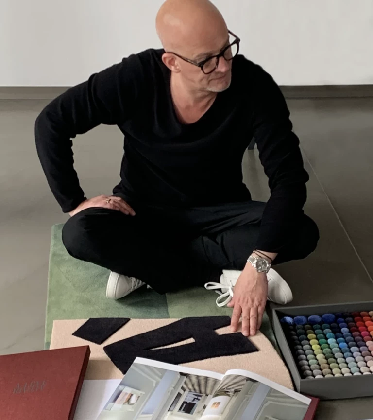 Olaf Kitzig sitzt auf dem Boden und zeigt auf einen Teppichentwurf, umgeben von Farbmustern und Designunterlagen.